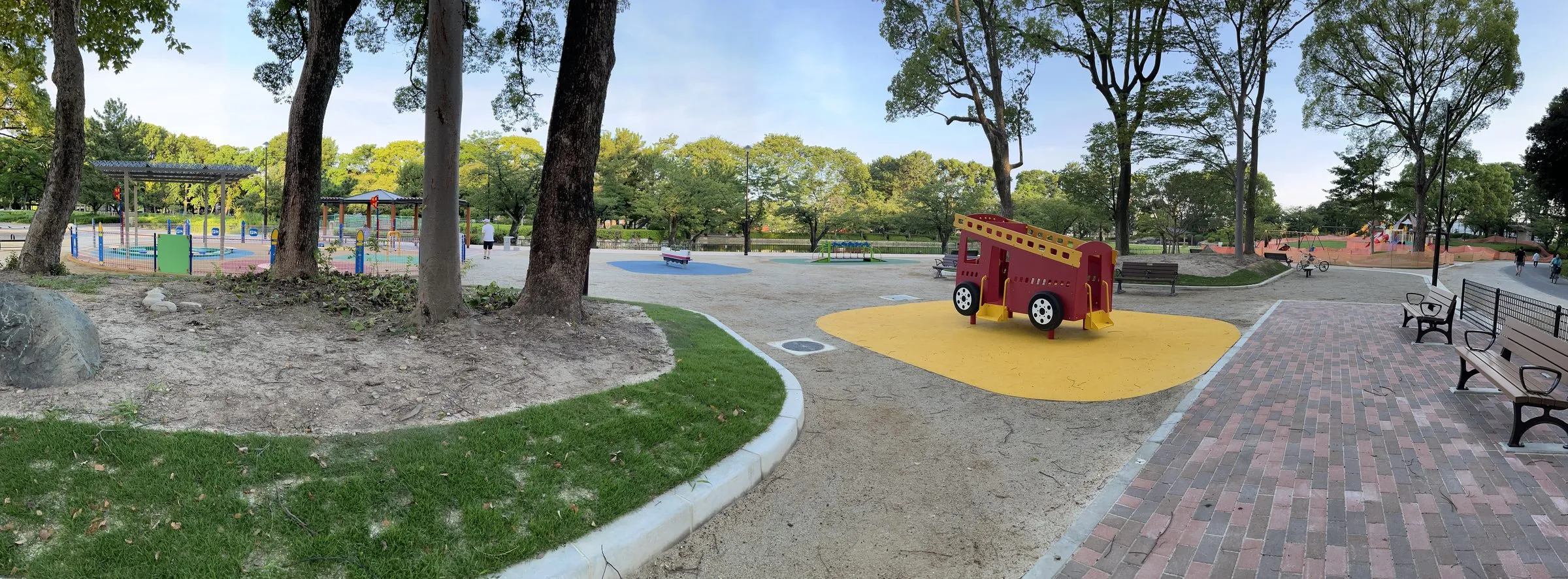 名城公園の子どもの広場の全景パノラマ。木陰とベンチ、赤い消防車の形をした遊具、奥に広がるカラフルな遊具エリア